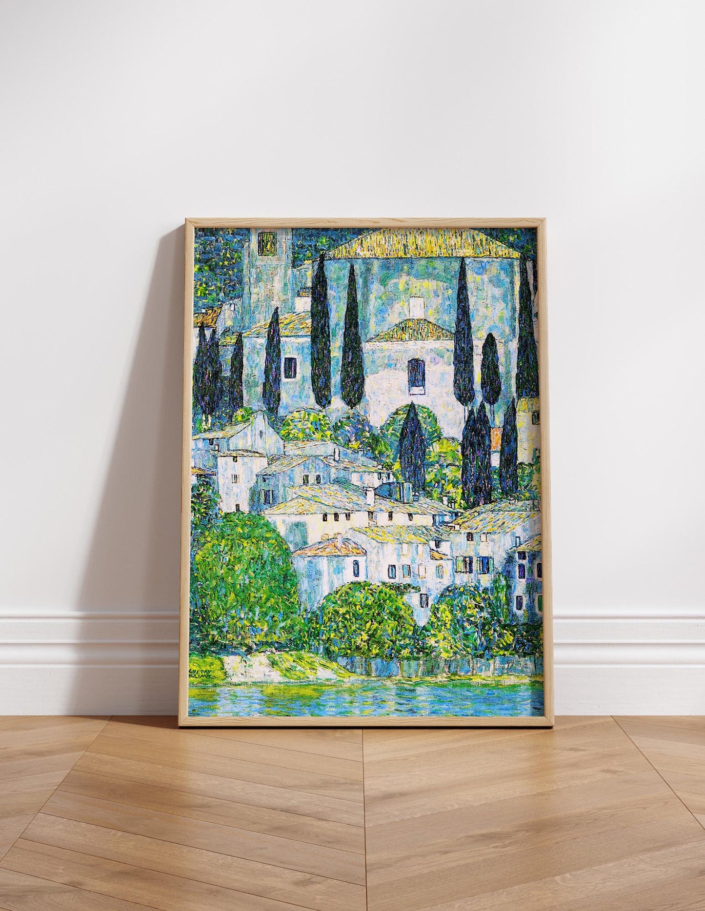 The image is a framed painting depicting a picturesque landscape with a village nestled among trees and a body of water in the foreground.